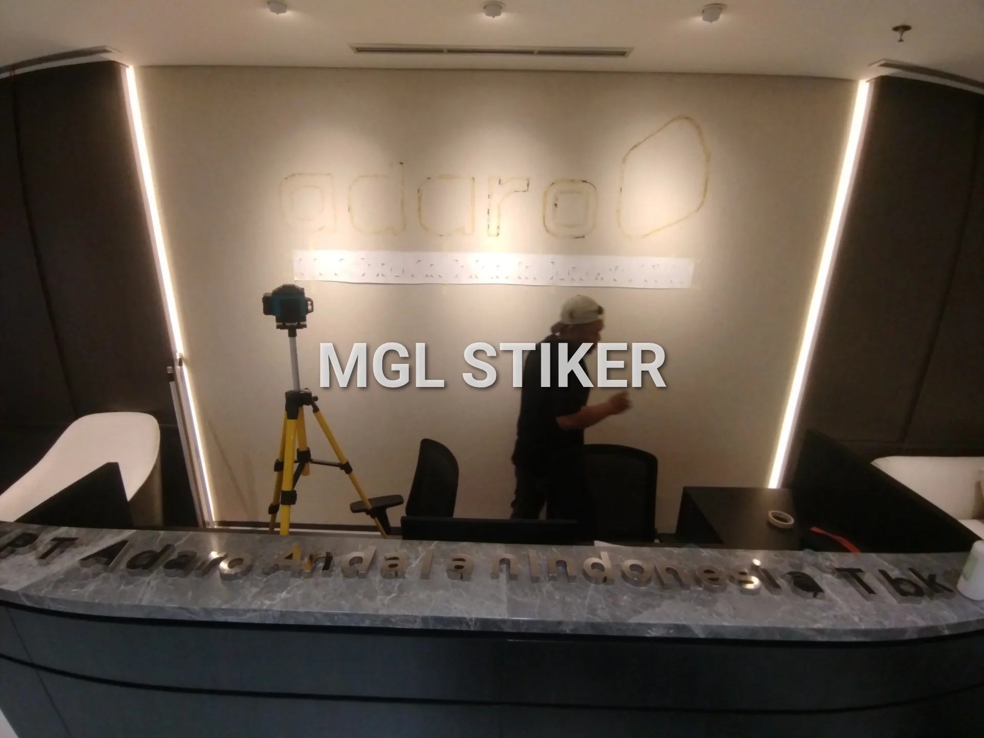 Pekerja MGL Stiker mempersiapkan pemasangan huruf timbul logo Adaro di dinding kantor, menggunakan panduan laser untuk presisi. MGL Stiker menyediakan solusi branding interior profesional seperti huruf timbul PT Adaro Andala Indonesia Tbk yang terlihat di meja resepsionis.