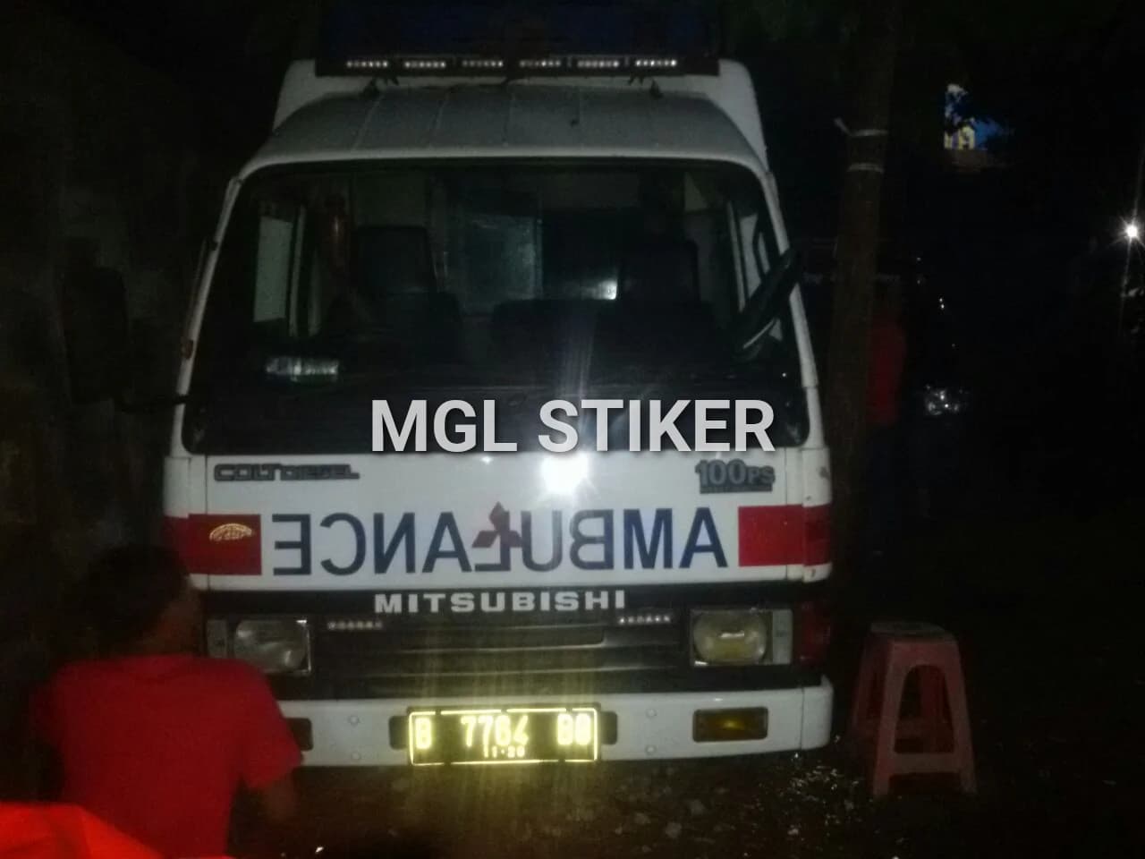 Depan mobil ambulans putih yang telah dilengkapi branding 'AMBULANS' terbalik menggunakan cutting sticker. Ini menunjukkan contoh layanan branding mobil profesional dari MGL Stiker untuk kendaraan operasional seperti ambulans Mitsubishi.