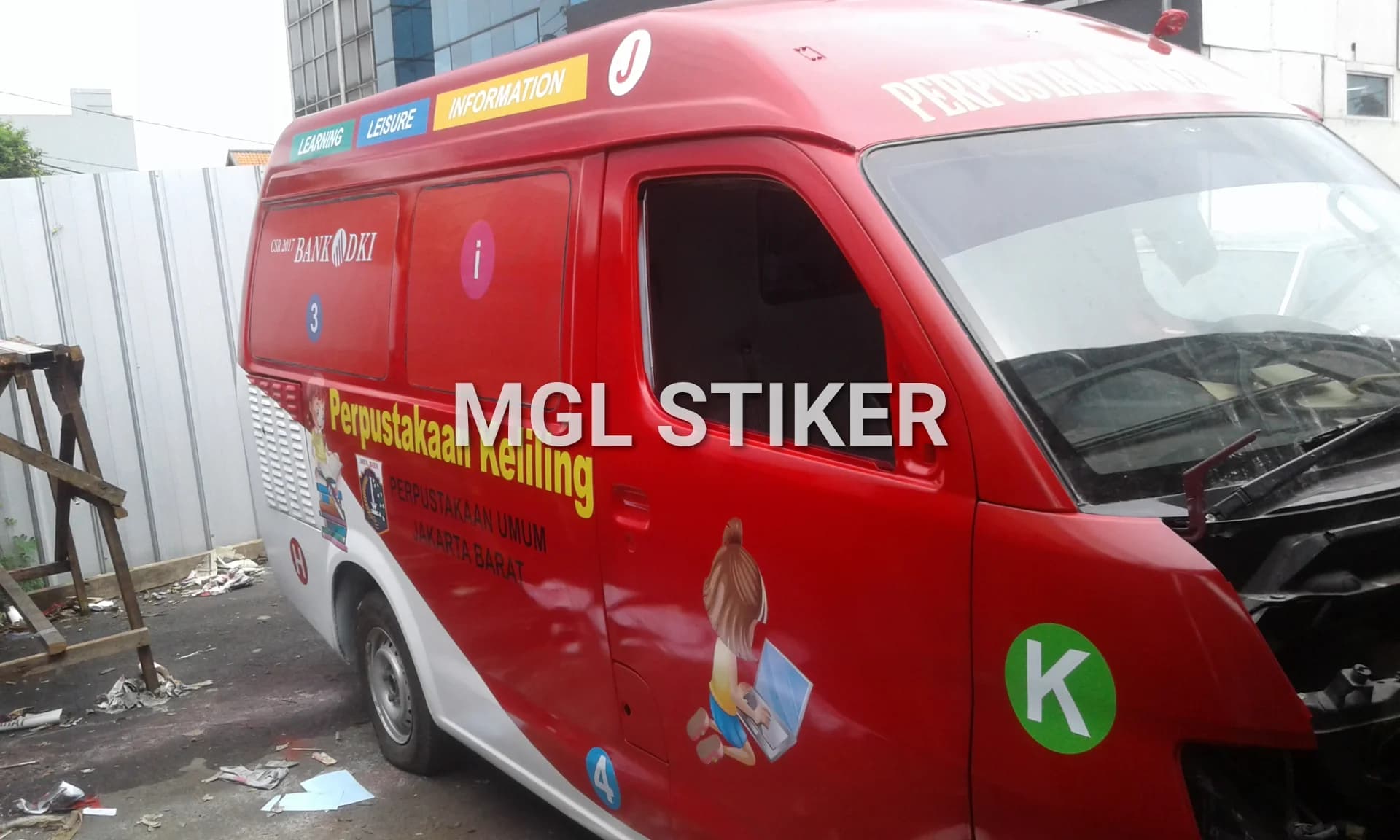 Branding mobil perpustakaan keliling warna merah dan putih dengan stiker logo Bank DKI dan Perpustakaan Umum Jakarta Barat. Desain ini menampilkan layanan edukasi 'Learning', 'Leisure', 'Information' serta ilustrasi anak dengan laptop, merupakan contoh proyek MGL Stiker.