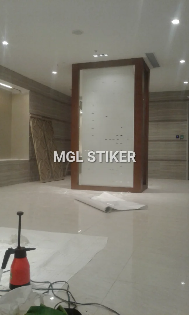 Gambar menunjukkan proses atau hasil aplikasi sticker sandblast motif cutting segitiga pada partisi kaca berbingkai kayu di ruangan modern. Terlihat roll sticker dan alat semprot di lantai, mengindikasikan proyek pemasangan. MGL Stiker adalah pilihan tepat untuk sticker sandblast dan cutting sticker berkualitas.